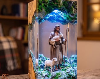 Könnte beinhalten: Eine rechteckige Lampe aus Harz und Holz mit einem beleuchteten Diorama einer Figur in braunem Gewand, die ein Lamm hält, während ein weiteres Lamm auf einem felsigen, moosigen Untergrund steht. Der obere Teil leuchtet blau, ein Schlüssel liegt davor.