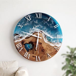 May include: A round wall clock featuring a natural wood grain base with a blue and white resin ocean wave design. The clock face displays white Roman numerals and thin white clock hands, creating a coastal aesthetic on the circular wood surface.