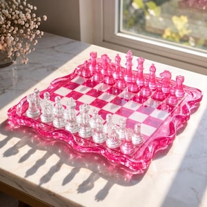 Op de afbeelding: Een vierkant schaakspel met een bord met afwisselend doorzichtige en doorschijnend roze vakjes. Het bord heeft een golvende, geschulpte rand in doorschijnend roze. Eén set schaakstukken is doorzichtig, de andere set is doorschijnend roze.