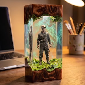May include: A rectangular resin lamp featuring a miniature miner figurine standing on a mossy base. The miner wears a hard hat with a light, a jumpsuit, and holds a pickaxe. Swirling green smoke effects are encased within the clear resin.