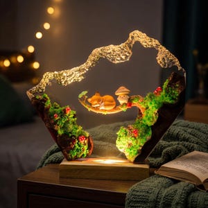 May include: A decorative resin lamp featuring a sleeping orange fox and a small mushroom character resting on a branch. The scene is set within a clear, mountain-shaped frame adorned with green moss, tiny red mushrooms, and a wooden base.