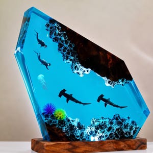 May include: A decorative resin sculpture featuring a deep blue underwater scene with two hammerhead sharks, two scuba divers, and a jellyfish. The piece incorporates dark wood elements at the top and bottom, with colorful sea anemones at the base.