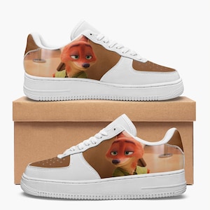 May include: A pair of white sneakers featuring a printed graphic of the animated fox character Nick Wilde from Zootopia. The shoes have white laces, a white sole, and brown panels on the toe and side, showcasing the character's orange fur and green shirt.