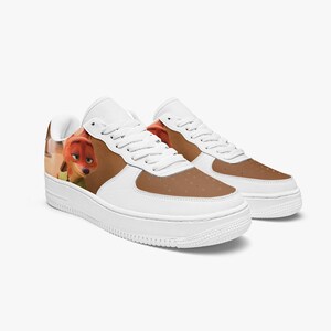 May include: A pair of white low-top sneakers featuring a brown panel on the toe box and side. The side of the shoe displays a printed graphic of a cartoon fox character with orange fur and green clothing, complete with white laces and a thick sole.