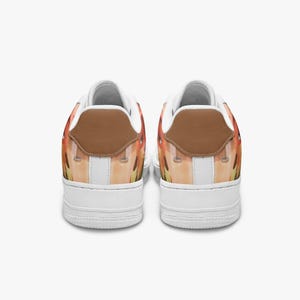 May include: A pair of low-top sneakers featuring a white rubber sole and a brown leather heel tab. The upper portion of the shoes displays a printed graphic with orange, yellow, and tan tones, complemented by white interior lining and white laces.