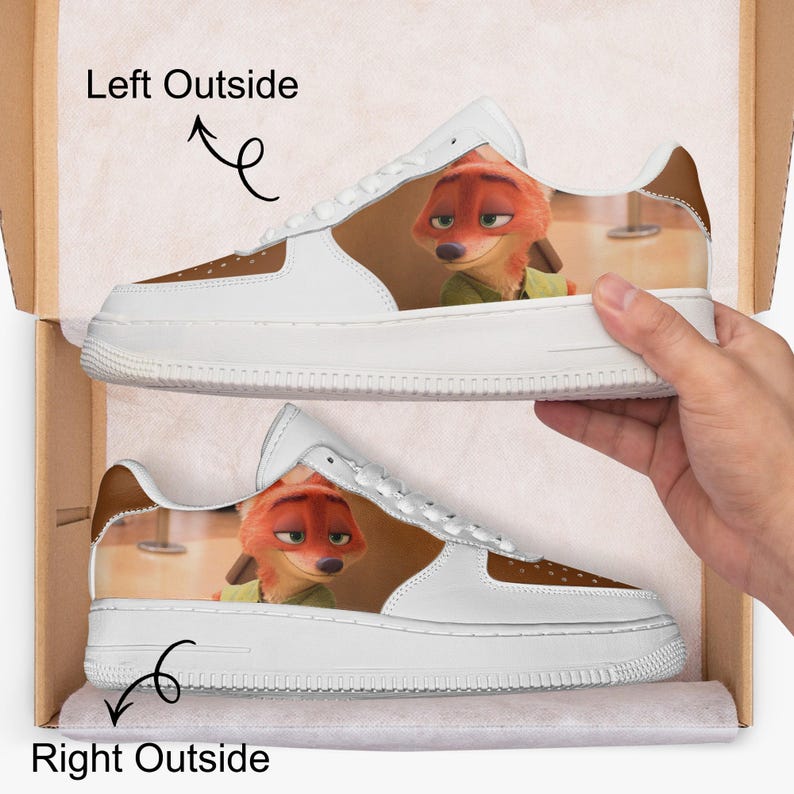 May include: A pair of white sneakers featuring a printed graphic of the orange fox character Nick Wilde from Zootopia on the side panels. The shoes have white laces, white soles, and brown accents near the toe and heel, presented in a cardboard box.