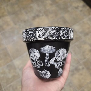 May include: A black plant pot featuring a white hand-painted mushroom design on the exterior. The rim of the pot is decorated with a repeating white circular floral pattern. The surface of the pot has a matte finish with textured white paint.