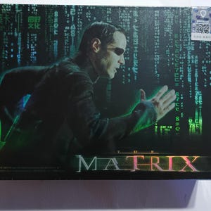 May include: A rectangular box featuring a graphic of a person in a black coat and dark sunglasses, set against a background of cascading green digital code. The title THE MATRIX is printed in large, metallic lettering at the bottom of the box.