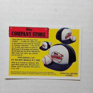 May include: A rectangular Topps Company Store special offer card featuring a yellow background with black text and two blue and white baseball caps. Each cap is adorned with a small plush baseball attached to the visor. The card includes promotional text detailing a mail-in offer for the caps, along with the Topps logo and mailing address in Westbury, New York.