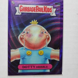 May include: A Garbage Pail Kids trading card featuring the character Dotty Dribble, a two-headed baby with a runny nose wearing a blue shirt with yellow smiley and sad face buttons, set against a vibrant, reflective purple holographic background.