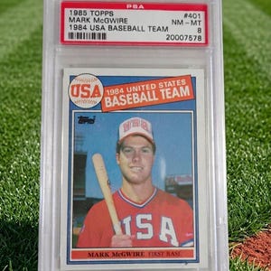 May include: A baseball card featuring Mark McGwire from the 1984 USA Baseball Team, graded by PSA. The card has a red and white design with the text "USA" and "1984 UNITED STATES BASEBALL TEAM". McGwire is shown holding a bat.