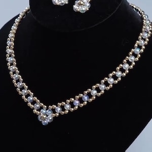 May include: A beaded necklace and matching pair of drop earrings featuring iridescent clear beads and small metallic gold beads. The necklace has a V-shaped center design with a flower-like cluster of beads, while the earrings mirror this pattern.