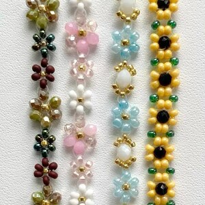 Peut inclure: Quatre brins de perles verticaux avec des motifs floraux. Le premier brin a des perles marron et vert olive. Le deuxième a des perles roses et blanches. Le troisième a des perles bleu clair et blanches. Le quatrième a des perles jaunes et noires.