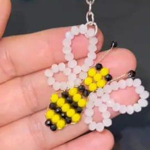 May include: A bee-shaped keyring crafted from small beads. The bee body features alternating yellow and black beads, while the wings are constructed from translucent white beads. The insect includes two black bead eyes and is attached to a silver metal ring.