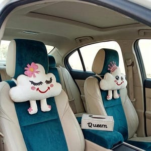 Peut inclure: Coussins de têtière de siège auto avec des motifs de nuages. Un nuage a une fleur rose, des cils et une bouche souriante. L'autre nuage a une couronne. Les sièges de voiture sont turquoise et beiges. Une boîte de mouchoirs avec le mot "Queen" est également visible.