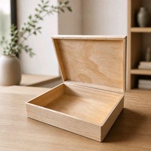 Puede incluir: Una caja rectangular de madera clara con una tapa abatible abierta. La caja presenta un acabado liso con veta de madera natural, paredes interiores visibles y una base plana. La construcción es sencilla y muestra el color madera clara.