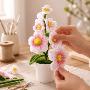 May include: A handcrafted pipe cleaner flower arrangement in a small white pot. The flowers feature white and pink petals with bright yellow centers, attached to green pipe cleaner stems. The base of the plant is covered in green moss.