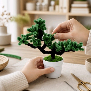 Op de afbeelding: Een kleine decoratieve boom gemaakt van donkerbruine pijpenragers met pluizig groen blad, geplant in een eenvoudige witte keramische pot gevuld met groen mos. De structuur bootst een bonsai-vorm na met een getextureerd, takachtig uiterlijk.