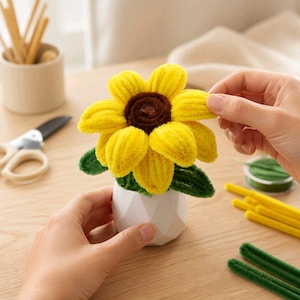 May include: A sunflower craft made from yellow and brown pipe cleaners, featuring a dark brown center and bright yellow petals. The flower is set in a small, white, geometric-shaped pot with green pipe cleaner leaves attached at the base.