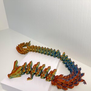 May include: A multi-colored articulated dragon figurine featuring a geometric, crystal-like texture. The body transitions in color from warm orange and yellow tones at the head to cool blue and purple hues along the long, segmented, flexible tail.