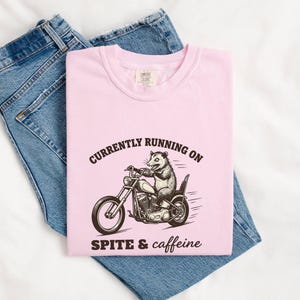 May include: A light pink t-shirt featuring a black graphic of an opossum riding a motorcycle. The text above the graphic reads "CURRENTLY RUNNING ON" and the text below reads "SPITE & caffeine". The shirt is folded over a pair of blue denim jeans.