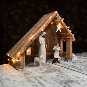 May include: A wooden nativity scene with a white star on top, featuring figurines of Mary, Joseph, and a baby Jesus, along with two sheep and a shepherd. The scene is illuminated with string lights.