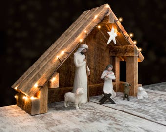 Può includere: Un presepe in legno con una stella bianca in cima, con figurine di Maria, Giuseppe e un bambino Gesù, insieme a due pecore e un pastore. La scena è illuminata da luci a catena.