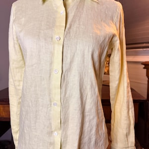 May include: A long-sleeved button-down shirt in a pale yellow color. The shirt features a classic collar, a front placket with five visible white buttons, and long sleeves with cuffs. The fabric has a visible textured weave throughout the garment.