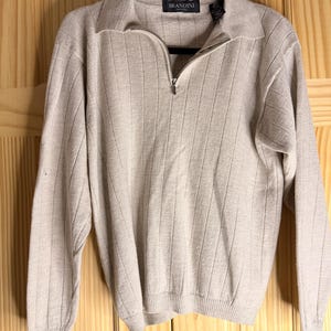 May include: A beige long-sleeve sweater with a vertical ribbed texture, a collared neckline, and a partial metal zipper closure. The garment features a black label at the inner neck with the text BRANDINI in white lettering, hanging on a black hanger.