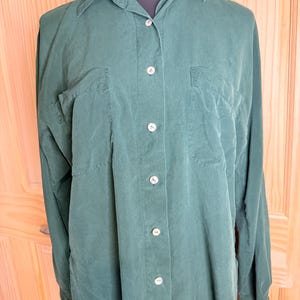 May include: A long-sleeved button-down shirt in a solid dark forest green color. The shirt features a classic collar, a front button placket with light-colored buttons, and two chest pockets with flaps. The fabric has a smooth, matte finish.