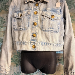May include: Light blue denim jacket with a classic collar, two chest pockets, and button closures. The jacket features a cropped design and is displayed on a black mannequin. The buttons are a light brown color.
