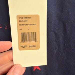 May include: A navy blue crewneck sweatshirt featuring a red star pattern. A cream-colored rectangular hang tag is attached, displaying the text STYLE: BLK8229CT2, COLOR: NAVY, CHAMPIONS CREWNECK, SIZE L, a barcode, and SUGG RETAIL PRICE $44.00.