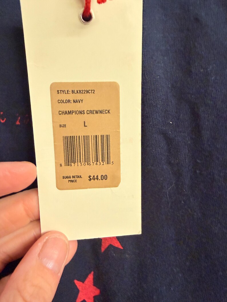 May include: A navy blue crewneck sweatshirt featuring a red star pattern. A cream-colored rectangular hang tag is attached, displaying the text STYLE: BLK8229CT2, COLOR: NAVY, CHAMPIONS CREWNECK, SIZE L, a barcode, and SUGG RETAIL PRICE $44.00.