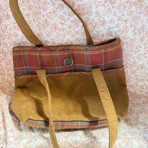 May include: A handbag featuring a tan suede-like lower section and a red, blue, and yellow plaid upper section. The bag has two tan shoulder straps and a central silver-toned circular button with embossed text on the front plaid panel.