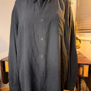 May include: A long-sleeved black corduroy shirt featuring a standard collar, a front button-down closure with matching black buttons, and a single patch pocket on the left chest. The shirt has buttoned cuffs and a vertical ribbed texture throughout.
