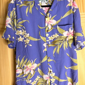 May include: A short-sleeved button-down shirt in a vibrant periwinkle blue featuring a repeating floral pattern of pink orchids and green palm leaves. The shirt includes a single chest pocket, a classic collar, and a Two Palms brand label.