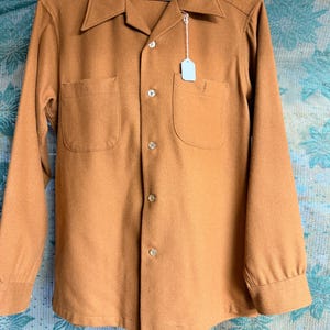 May include: A long-sleeved, button-down shirt in a warm, golden-brown hue. The shirt features two front pockets, a classic collar, and a tag attached. The fabric appears to be a soft, wool-like material, perfect for cooler weather.