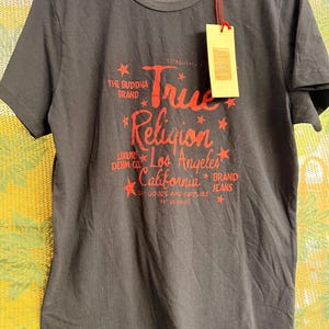 May include: A charcoal gray short-sleeve t-shirt featuring red graphic text that reads True Religion Los Angeles California, The Buddha Brand, Luxury Denim Co., and Brand Jeans, accented with small red stars and a hanging beige paper tag.