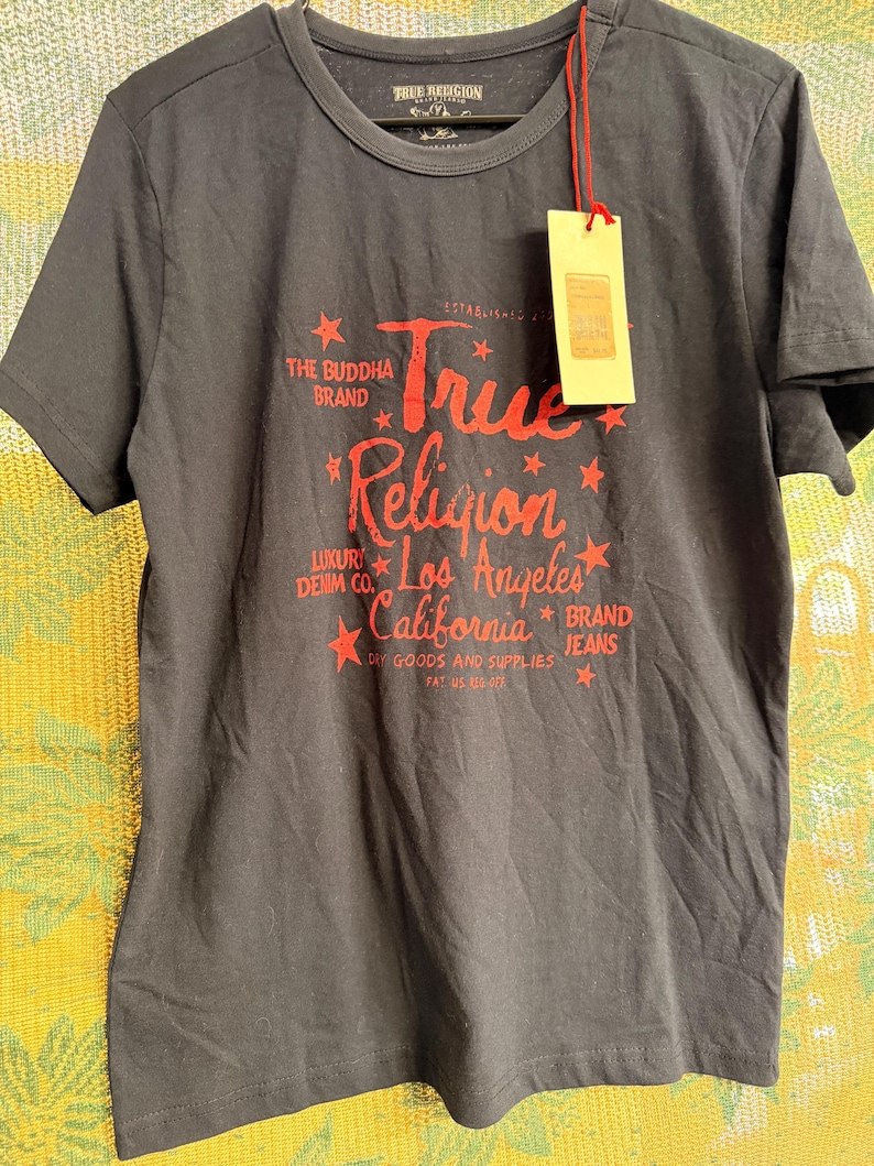 May include: A charcoal gray short-sleeve t-shirt featuring red graphic text that reads True Religion Los Angeles California, The Buddha Brand, Luxury Denim Co., and Brand Jeans, accented with small red stars and a hanging beige paper tag.