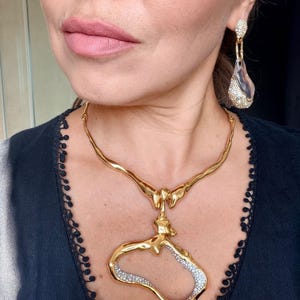 Può includere: Una collana rigida in metallo dorato con un pendente organico e asimmetrico, caratterizzato da un'apertura centrale e cristalli incastonati. L'orecchino abbinato è composto da un perno tempestato di cristalli e una forma in resina trasparente.