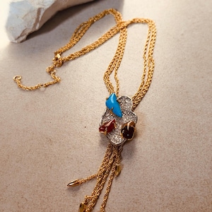 Può includere: Una collana bolo dorata con un ciondolo centrale a forma di nuvola incastonato di cristalli chiari. Il ciondolo è accentuato da una pietra blu brillante, una rossa scura e una marrone scuro. Tre catene dorate pendono dalla base.