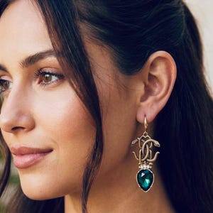 Può includere: Un orecchino pendente color oro caratterizzato da un motivo a serpenti intrecciati e testurizzati nella parte superiore, accentuato da una fila di piccoli cristalli trasparenti e rifinito con una grande gemma sfaccettata verde smeraldo.