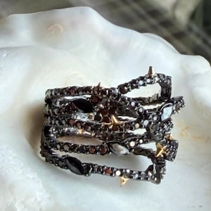 May include: A multi-band ring featuring a dark, textured finish with embedded black stones and small, pointed gold-toned accents. The design consists of several interconnected, winding bands that create an open, layered structure.