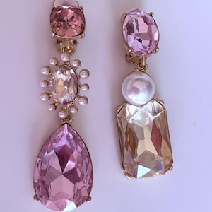 Peut inclure: Une paire de boucles d'oreilles pendantes dépareillées avec des montures dorées. L'une présente un cristal ovale rose, un cristal ovale central entouré de petites perles blanches et une grande goutte rose. L'autre a un cristal ovale rose, une perle blanche et un grand cristal rectangulaire couleur champagne.