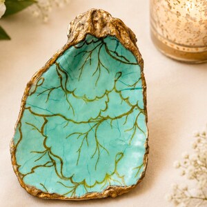 May include: A decorative oyster shell dish featuring a vibrant turquoise interior with hand-painted gold branch-like patterns. The exterior edges of the shell are finished with a textured gold metallic trim, creating a distinct contrast against the smooth, glossy turquoise surface of the concave shell.