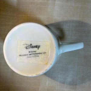 May include: A white ceramic mug base featuring a rectangular sticker with the Disney logo. The text on the sticker reads: Disney, Microwave and Dishwasher Safe, Made in China. The mug has a smooth white handle extending from the side of the circular base.