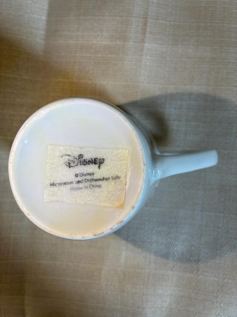 May include: A white ceramic mug base featuring a rectangular sticker with the Disney logo. The text on the sticker reads: Disney, Microwave and Dishwasher Safe, Made in China. The mug has a smooth white handle extending from the side of the circular base.