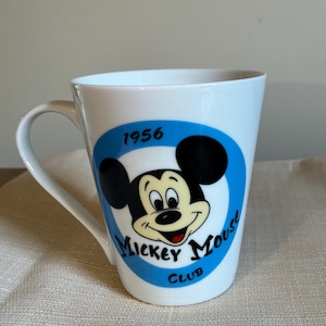 May include: A white ceramic mug featuring a blue circular graphic with the black and cream-colored face of Mickey Mouse. The text 1956 is printed above the character, and the words Mickey Mouse Club are printed in black below the character.
