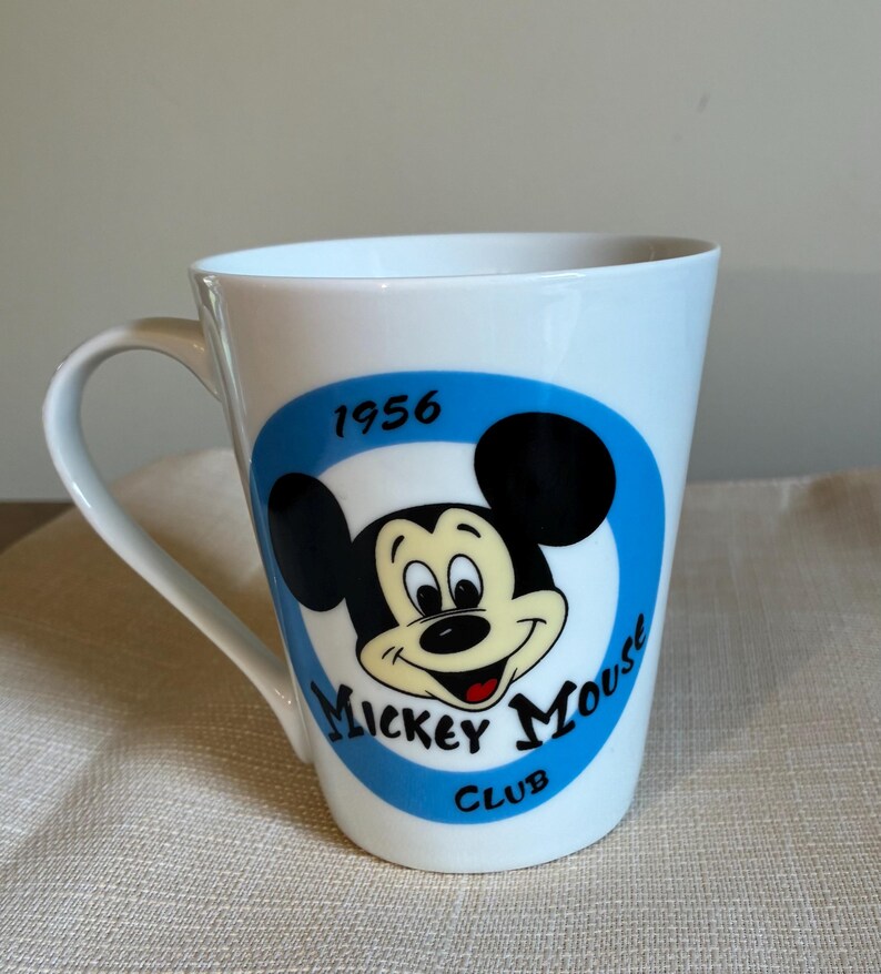 May include: A white ceramic mug featuring a blue circular graphic with the black and cream-colored face of Mickey Mouse. The text 1956 is printed above the character, and the words Mickey Mouse Club are printed in black below the character.