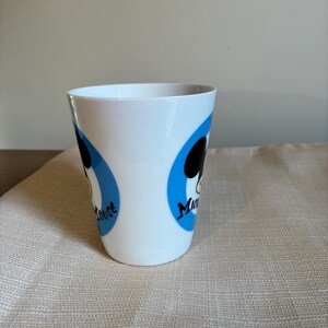 May include: A white ceramic mug featuring two blue circular graphics, each containing a black silhouette of Mickey Mouse's head and the text "Mickey Mouse" in black script. The mug has a smooth, tapered cylindrical shape with a wide rim.